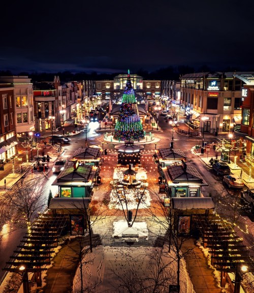A festive night scene featuring a brightly lit Christmas tree in a bustling plaza, surrounded by shops and holiday lights.