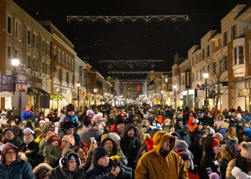A large crowd gathers on a festive street at night, adorned with holiday lights and decorations.