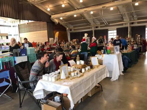 A bustling indoor market with vendors and shoppers, featuring tables of handmade goods and festive decorations.