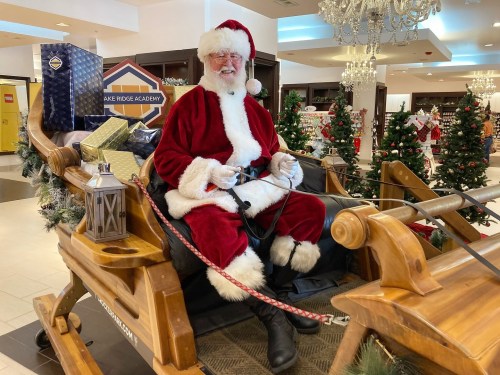 Santa Claus sits in a wooden sleigh surrounded by Christmas decorations and gifts in a festive indoor setting.