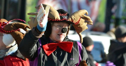 A woman in a festive reindeer costume waves, wearing antlers and a red bow tie, smiling at a parade.