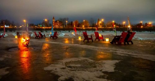 Red chairs by a frozen beach at night, with a fire pit and glowing lanterns illuminating the snowy landscape.