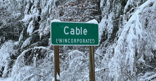 A green sign reading "Cable UNINCORPORATED" surrounded by snow-covered trees.