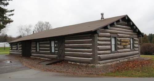 A rustic log cabin with a sloped roof, set in a grassy area under a cloudy sky.