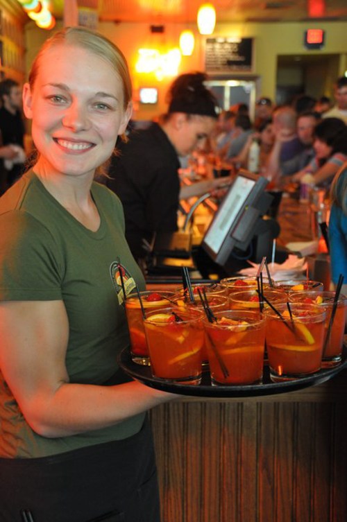 A smiling waitress holds a tray of colorful drinks at a busy bar, with patrons in the background.