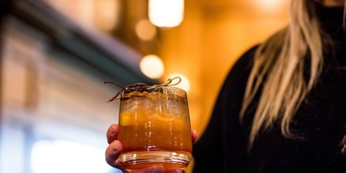 A person holding a cocktail garnished with a cherry and citrus peel, with warm lighting in the background.