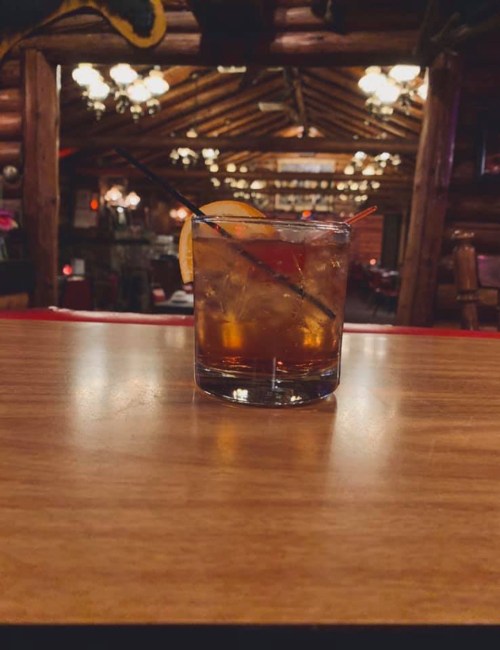 A glass of whiskey with ice and a lemon slice sits on a wooden table in a cozy, rustic cabin interior.