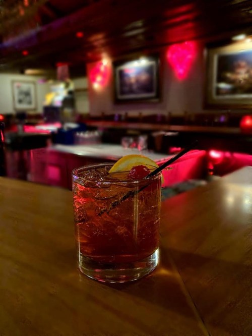 A cocktail with a lemon slice and cherry sits on a bar, illuminated by soft red lights in a cozy setting.