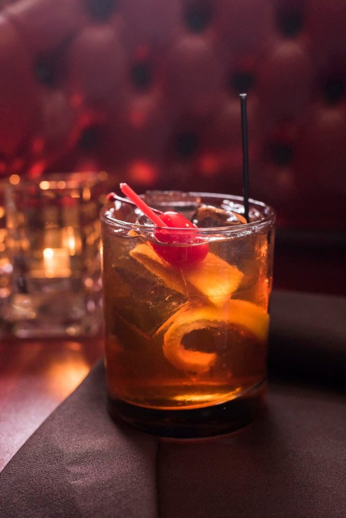 A cocktail in a glass with ice, garnished with a cherry and citrus slices, set on a dark table.