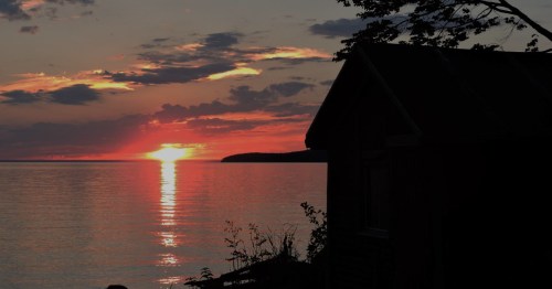 Sunset over calm water, with a silhouette of a building and trees in the foreground.