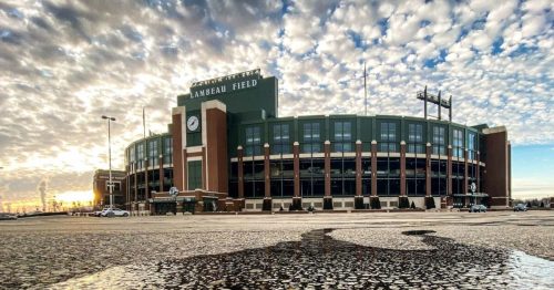 Lambeau Field at sunset, showcasing its iconic architecture against a cloudy sky and reflecting puddles in the foreground.
