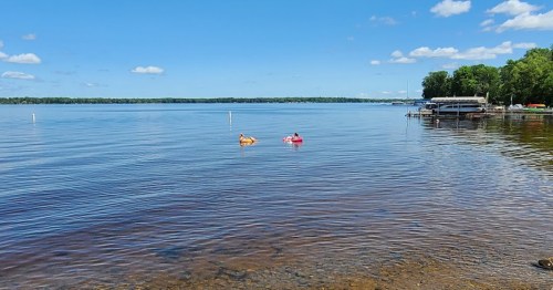 Two people float on colorful rafts in a calm lake under a clear blue sky, with trees and a dock in the background.