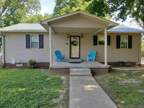 A cozy house with a front porch, two blue chairs, and a walkway surrounded by green grass and trees.