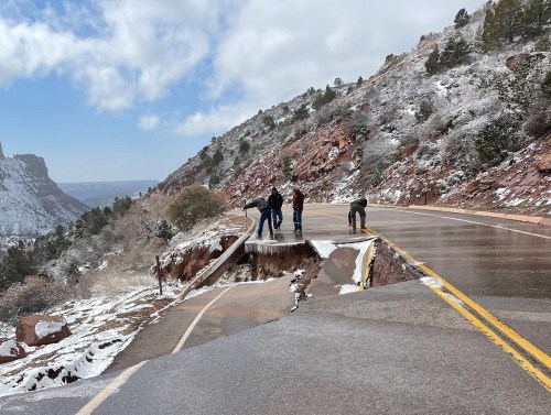 A group of people examines a damaged road with a large section collapsed, surrounded by snow-covered mountains.