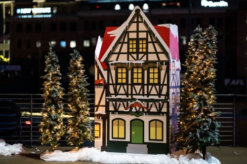 A festive, oversized decorative house with a snowy base, flanked by two Christmas trees, illuminated at night.