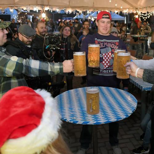 A group of people cheers with large beer mugs at a festive outdoor gathering, with holiday decorations in the background.