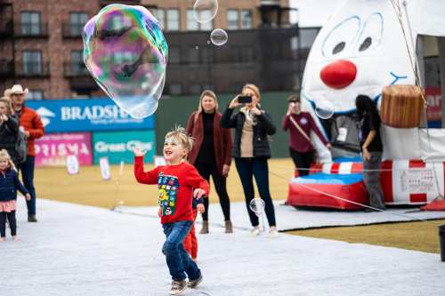 A child in a red shirt joyfully chases a large bubble at an outdoor event, with people and a festive backdrop behind them.