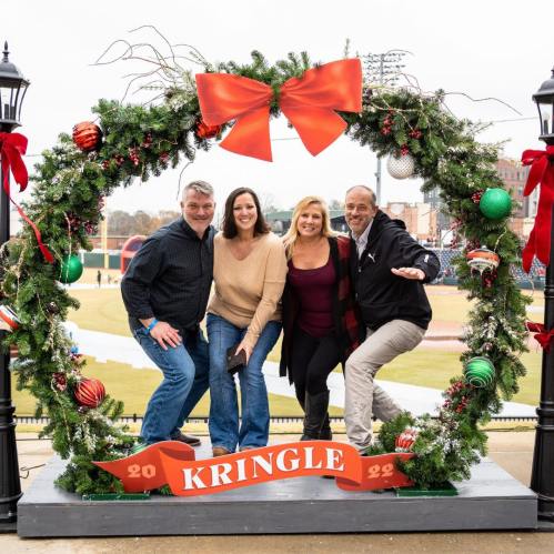 A group of four friends poses cheerfully in a festive holiday wreath with decorations and a large red bow.