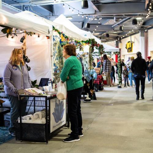 A bustling market scene with vendors in tents, shoppers browsing, and festive decorations in a spacious indoor area.