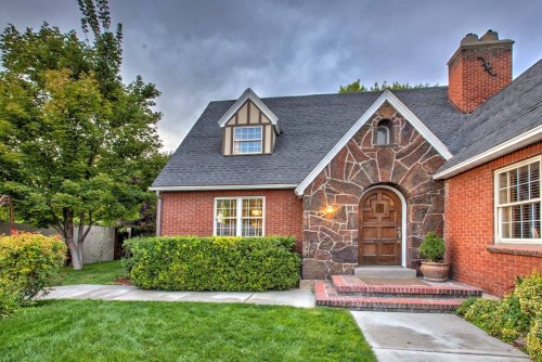 A charming brick house with a stone accent, surrounded by greenery and a well-maintained lawn under a cloudy sky.