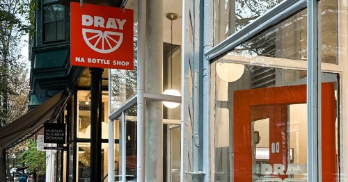 Exterior of Dray NA Bottle Shop, featuring a bright orange sign and large windows, with trees in the background.