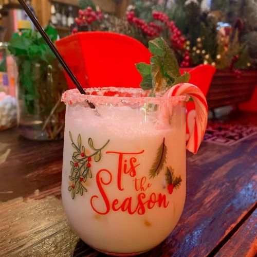 A festive drink in a glass with "Tis the Season" design, garnished with a candy cane and surrounded by holiday decor.