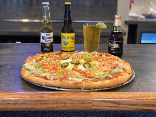 A large pizza topped with lettuce, tomatoes, jalapeños, and cheese, accompanied by three bottles of beer on a table.