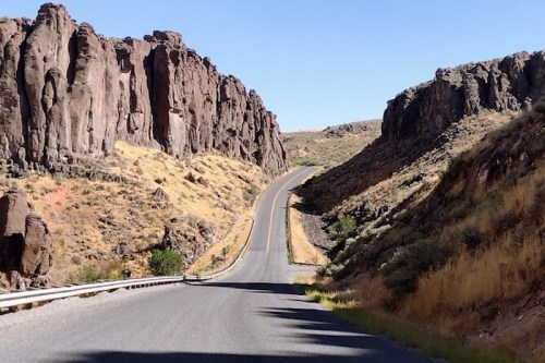 A winding road cuts through rocky cliffs and dry hills under a clear blue sky.