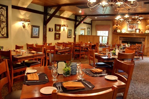 Cozy restaurant interior with wooden tables, chairs, and elegant table settings, featuring warm lighting and decor.