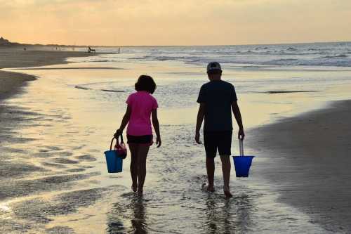 Two children walk along a beach at sunset, carrying blue buckets and wading through the shallow water.