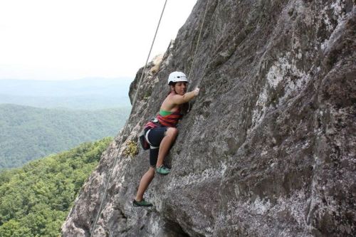 A climber in a helmet ascends a rocky cliff with a scenic mountain backdrop.