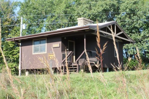 A small brown cabin with a stone chimney, surrounded by tall grass and trees in the background.