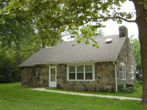 A stone cottage with a sloped roof, surrounded by green grass and trees.
