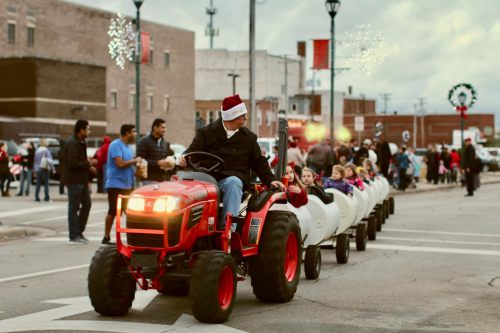 A man in a Santa hat drives a tractor pulling a festive train of children through a holiday parade.