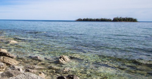 A clear blue lake with rocky shoreline and a small island in the distance under a light blue sky.