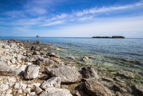 A rocky shoreline with clear water, leading to a distant island under a blue sky with scattered clouds.