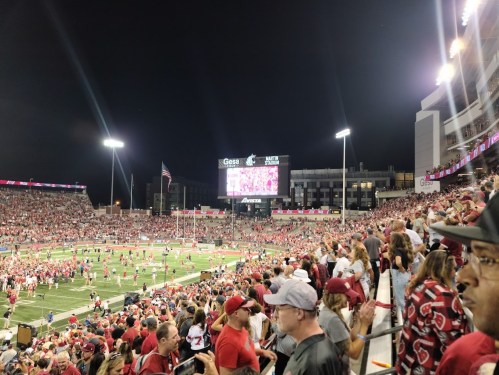 A crowded stadium at night, filled with fans in red, celebrating on the field with bright lights and a large screen.