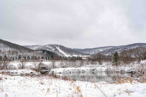 A serene winter landscape featuring a snowy field, a calm pond, and distant hills under a cloudy sky.