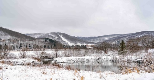A serene winter landscape featuring snow-covered hills, a calm river, and bare trees under a cloudy sky.