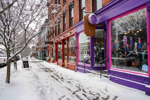 Colorful storefronts line a snowy street, with trees and a clear winter sky in the background.