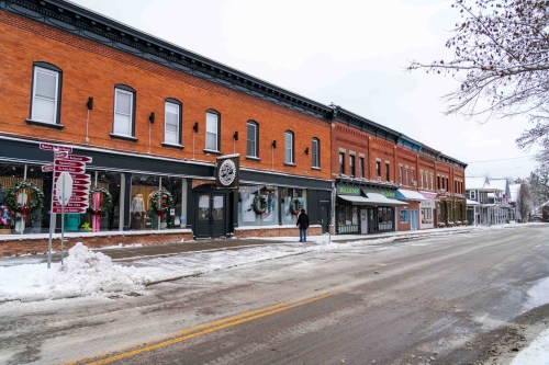 A snowy street lined with brick buildings, featuring shops decorated for the holidays and a person walking by.