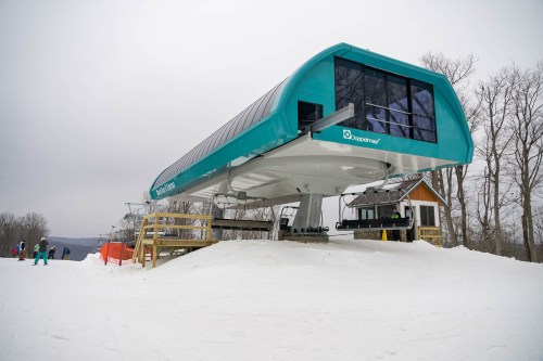 A modern ski lift station on a snowy slope, surrounded by trees and skiers in the background.
