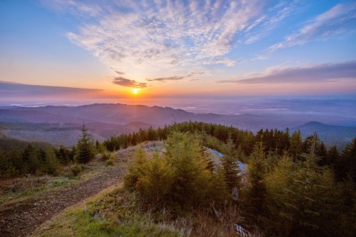 A scenic view of a sunrise over rolling hills and forests, with a colorful sky and mist in the valleys below.
