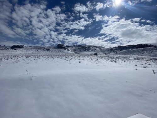 A snowy landscape under a bright sky with scattered clouds and distant hills. Sunlight glimmers on the snow.