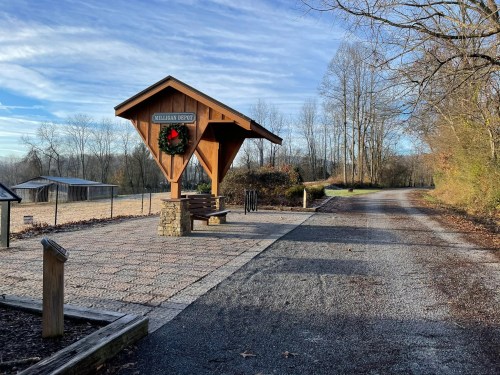 A wooden shelter with a wreath stands by a gravel path surrounded by trees and open fields under a clear sky.
