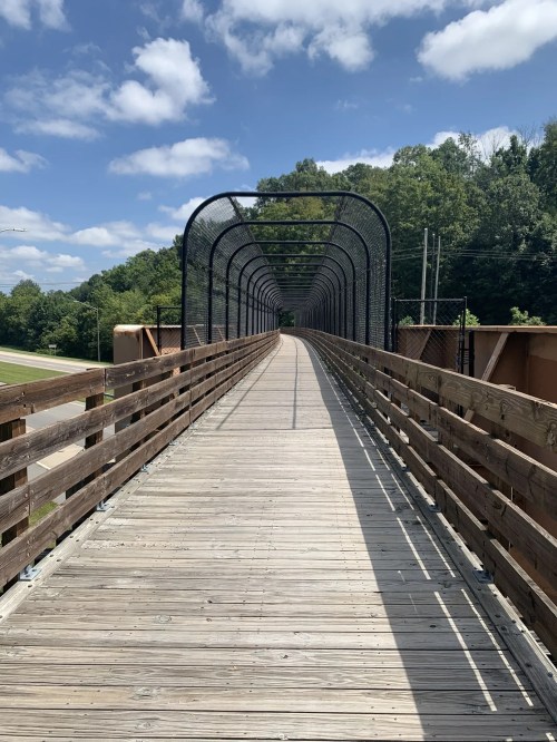 A long wooden bridge with a curved metal frame, surrounded by greenery and a blue sky with clouds.