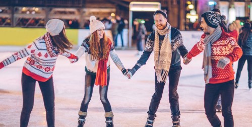 Four friends ice skating together, wearing festive sweaters and smiling, on a rink with colorful lights in the background.