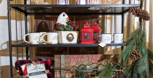A metal shelf displays festive decor, including mugs, a lantern, a wreath, and pinecones, with a cozy blanket below.