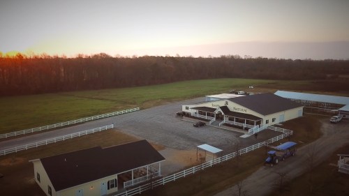 Aerial view of a large building on a farm, surrounded by green fields and trees, with a sunset in the background.