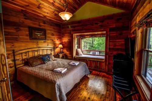 Cozy wooden bedroom with a double bed, soft linens, and a window seat overlooking nature. Warm, inviting atmosphere.